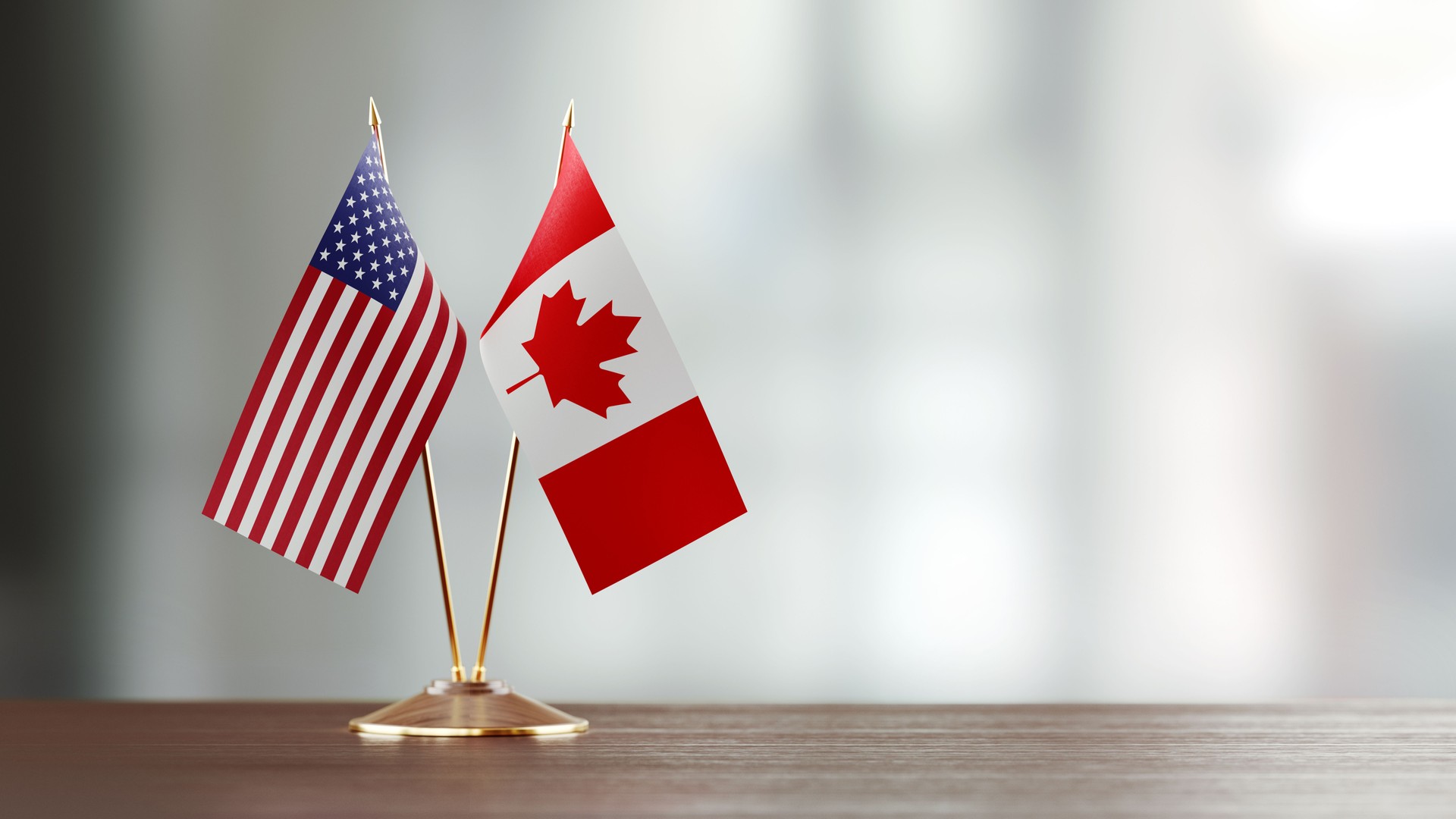 American And Canadian Flag Pair On A Desk Over Defocused Background